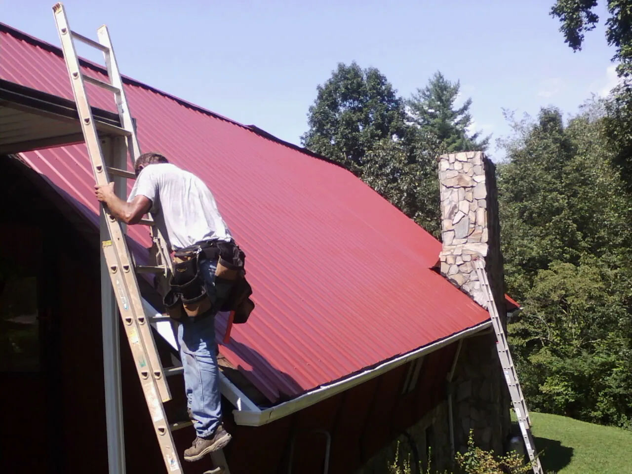 Roofer on a ladder beside a red metal roof and stone chimney — Soffit & Fascia Repair by Roofing Brothers in Sweet